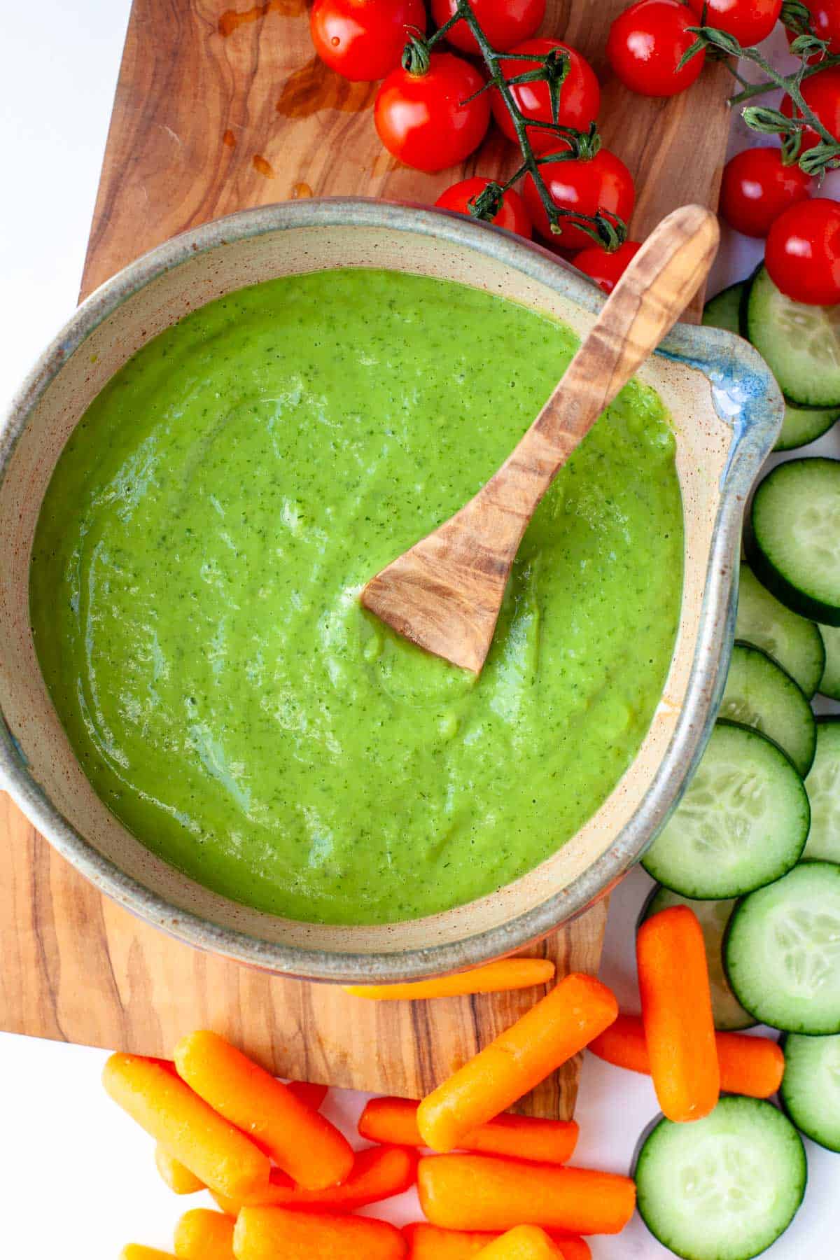 Vegan Green Goddess Dressing in serving bowl with wooden spoon on top of wooden cutting board and next to fresh veggies for dipping.