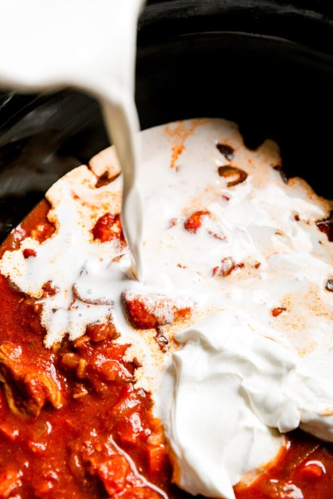 Cream being poured on butter chicken ingredients in a crock pot. 