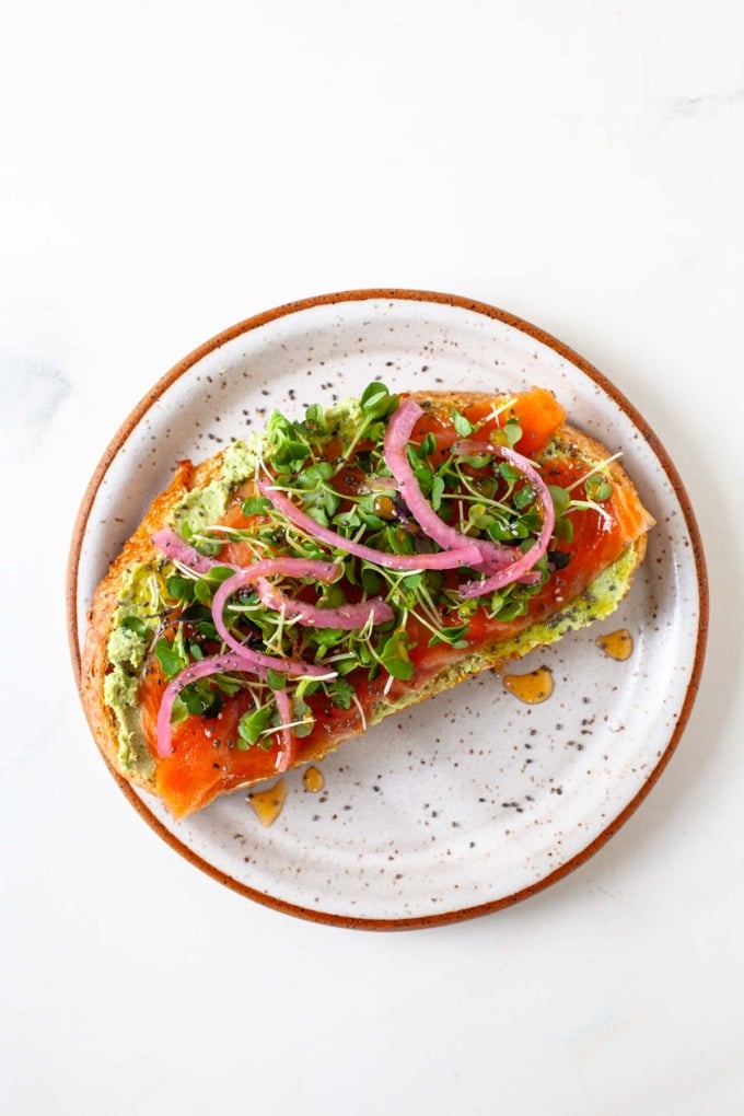 slice of toast with edamame spread, smoked salmon, microgreens, pickled onions and honey on plate