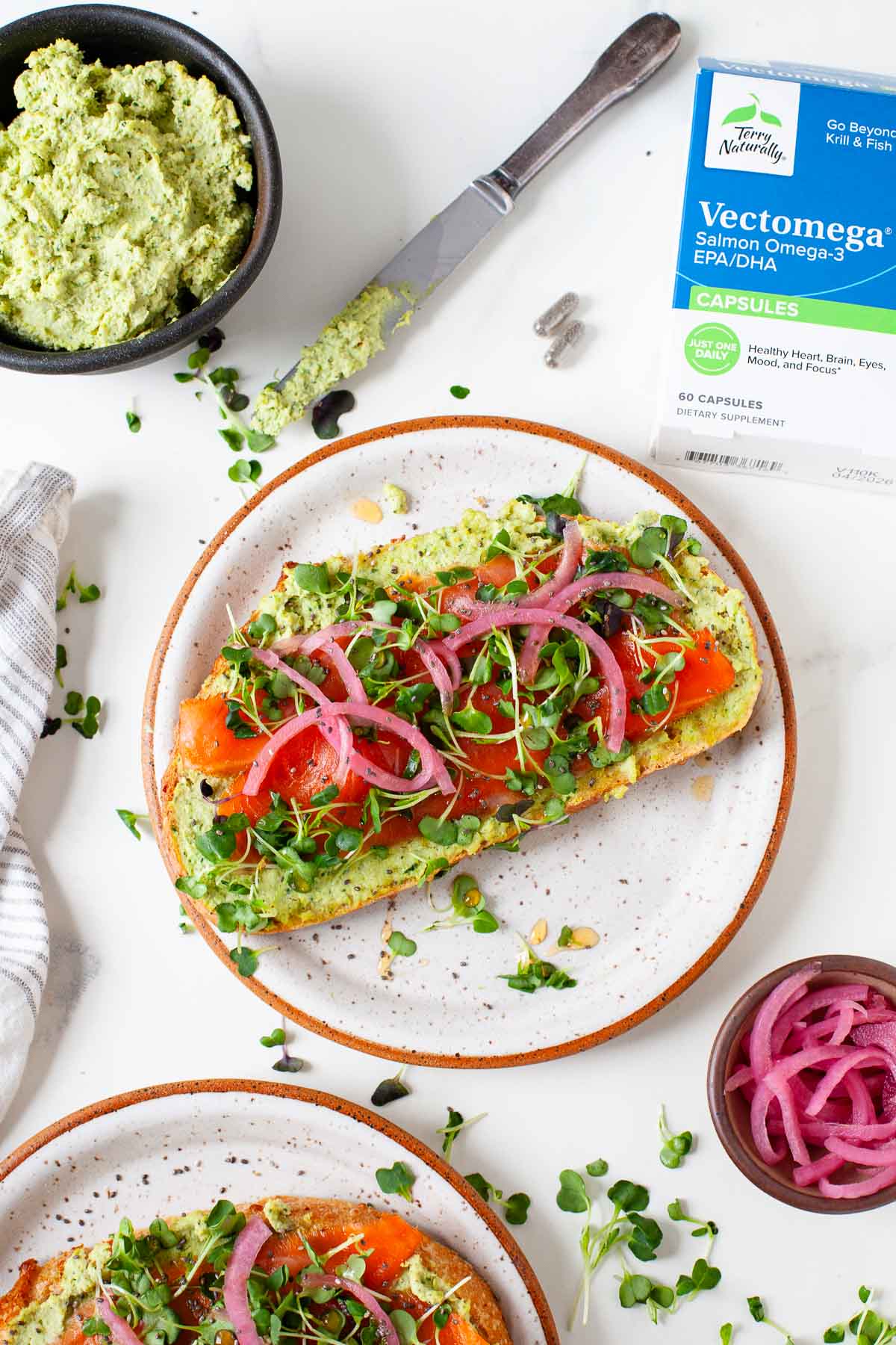 omega 3 supplement near two plates with smoked salmon toast and bowls with edamame spread and pickled onions